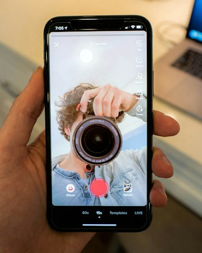 Person, die sich selbst im Smartphone betrachtet. Das Gesicht ist allerdings nicht zu erkennen, weil mitten auf dem Smartphone-Bildschirm ein großes Kamera-Objektiv angebracht ist.