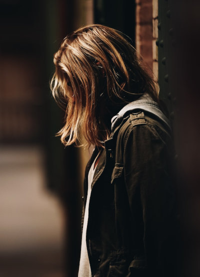 Eine Frau ist von der Seite zu sehen. Sie sieht nach unten und ihre Haare fallen ins Gesicht, so dass sie nicht zu erkennen ist. Die Szenerie ist dunkel und einsam.