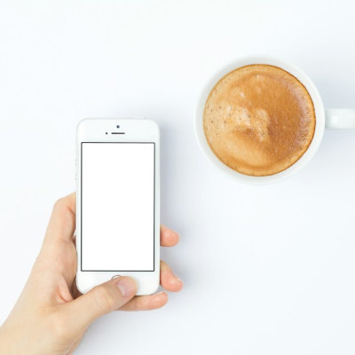 Eine Hand hält ein weißes Smartphone mit weißem Bildschirm, daneben eine weiße Tasse mit braunem Cappuccino, alles sehr schick.