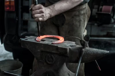 Schmied in Nahaufnahme bei der Arbeit, nur seine linke Hand mit einem Teil des Unterarms ist zu sehen. Er hämmert ein glühendes Hufeisen. Das Bild ist in gedeckten Grau- und Brauntönen gehalten, nur das Hufeisen glüht kräftig orange.